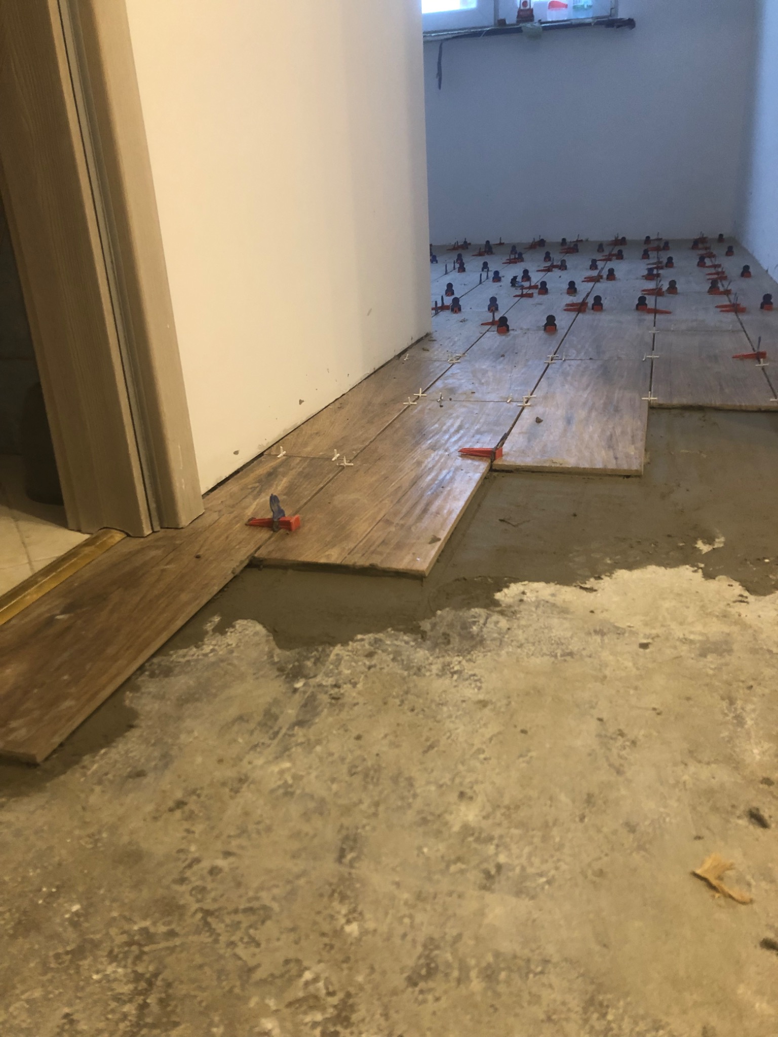 Podłoga w trakcie układania płytek imitujących drewno, z wykorzystaniem systemu poziomowania płytek, widoczne nierówności podłoża i świeży klej.