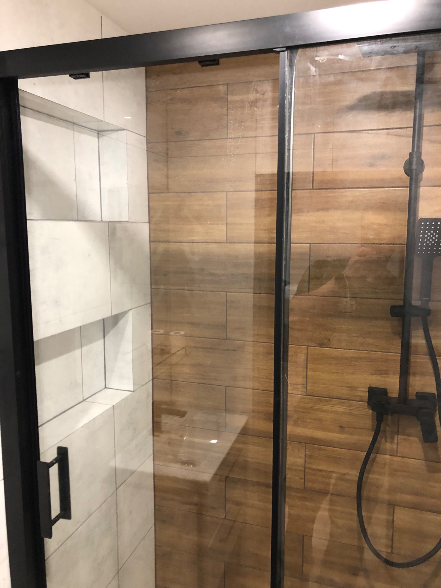 Kabina prysznicowa z przesuwnymi drzwiami, wykończona płytkami imitującymi drewno i białymi płytkami z wnękami, czarna armatura.