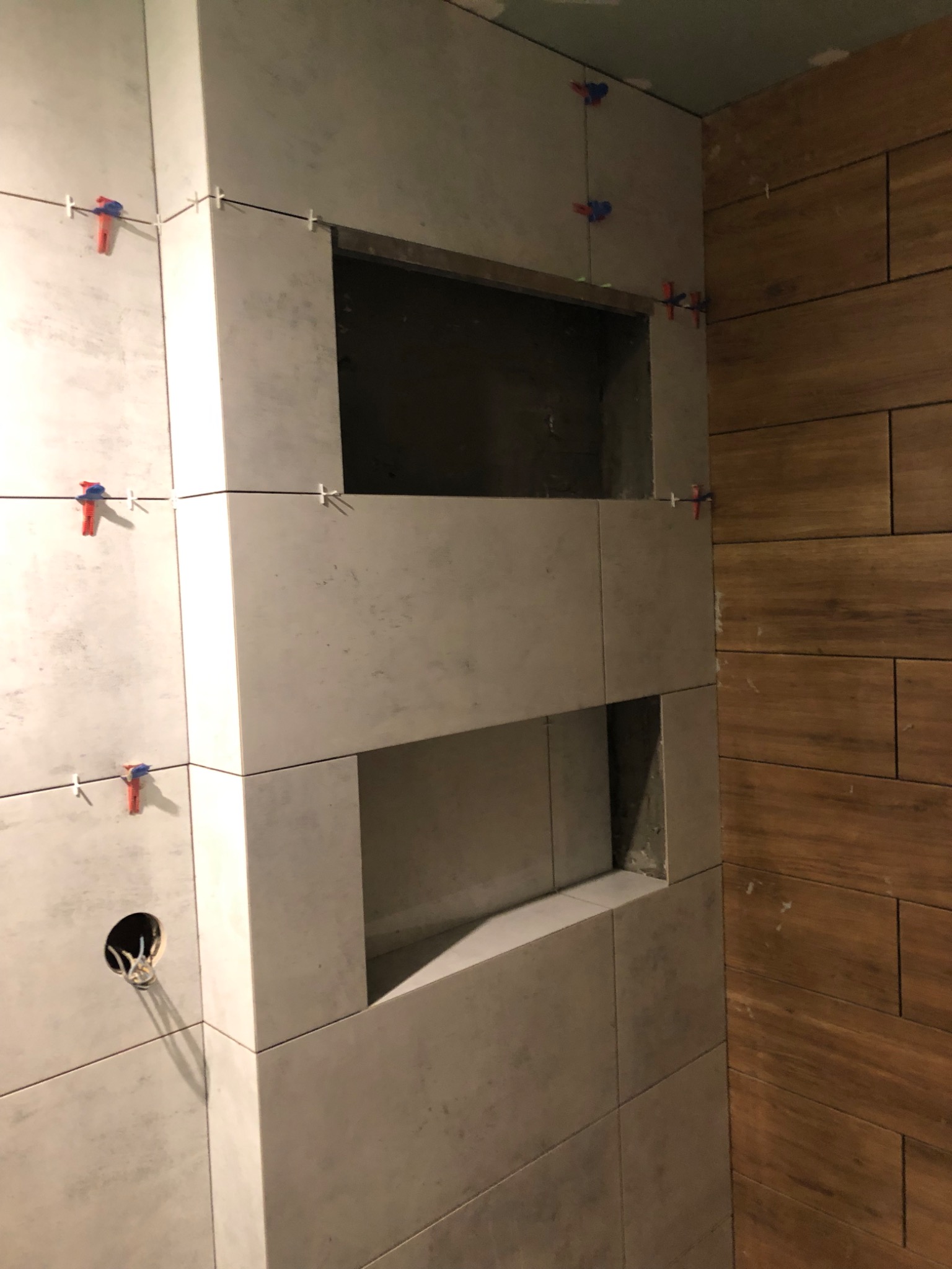 Ściana w trakcie układania płytek z wnękami, widoczne krzyżyki dystansowe i otwór na instalację elektryczną, obok fragment ściany wykończonej drewnopodobnymi płytkami.