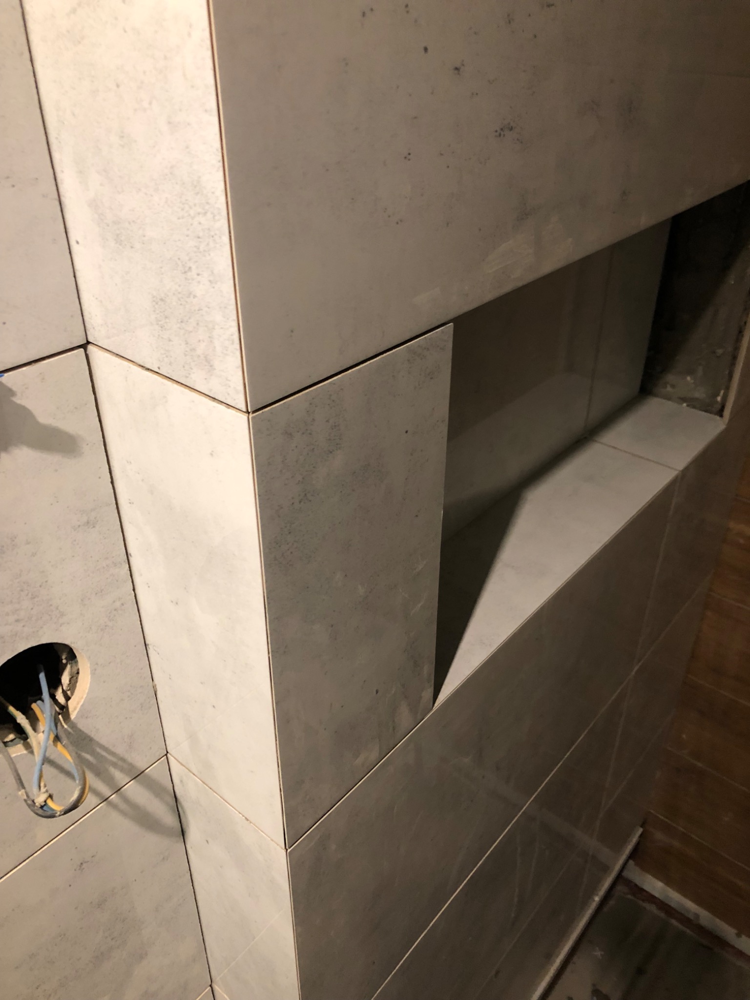 Ściana łazienki z szarymi, matowymi płytkami ceramicznymi, wnęka w ścianie wykończona tymi samymi płytkami, widoczne przewody elektryczne wystające z otworu w ścianie.