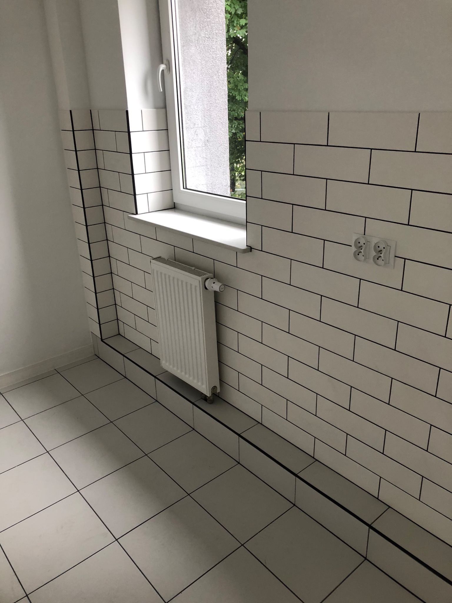 Białe, prostokątne płytki z czarną fugą na ścianie i podłodze w łazience, grzejnik pod oknem, gniazdka elektryczne na ścianie.