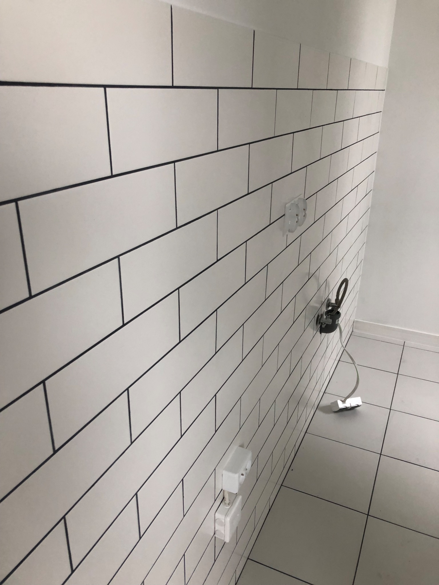 Ściana wyłożona białymi, prostokątnymi płytkami z ciemną fugą, widoczne gniazdka elektryczne i przewody, świeżo po montażu.