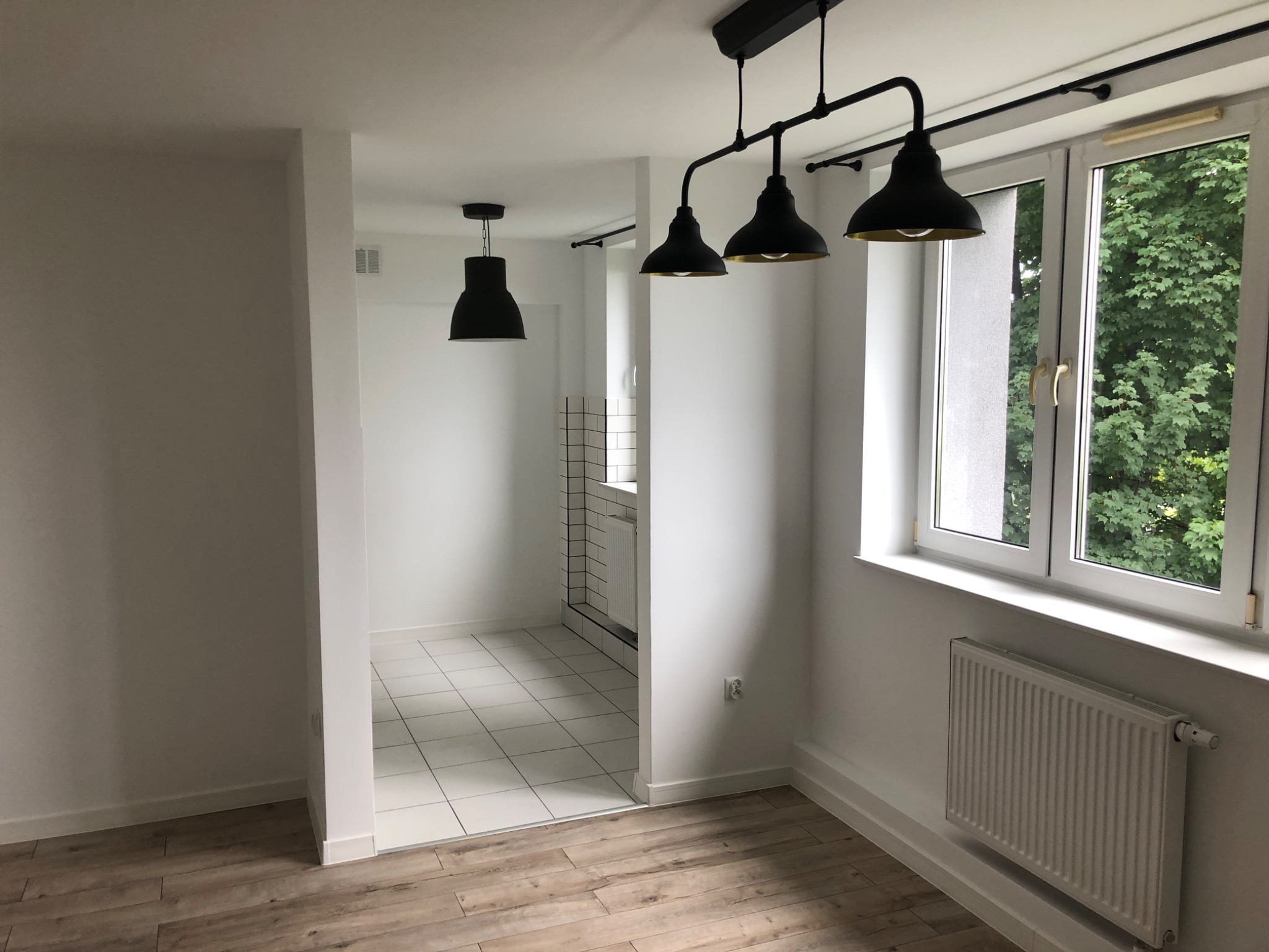 Pomalowane na biało pomieszczenie z drewnopodobną podłogą, trzema czarnymi lampami wiszącymi w stylu industrialnym i widokiem na zieleń za oknem. W tle widoczna biała łazienka z białymi płytkami...