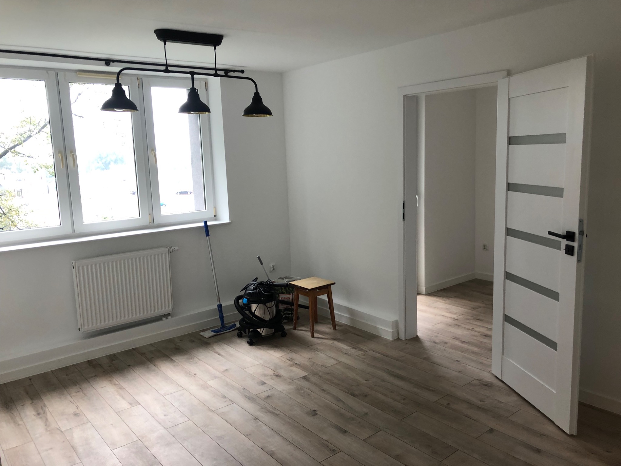 Pusty, jasny pokój z drewnopodobną podłogą, białymi ścianami i grzejnikiem pod oknem, widoczny odkurzacz przemysłowy, drabinka i mop, drzwi wewnętrzne z przeszkleniami.