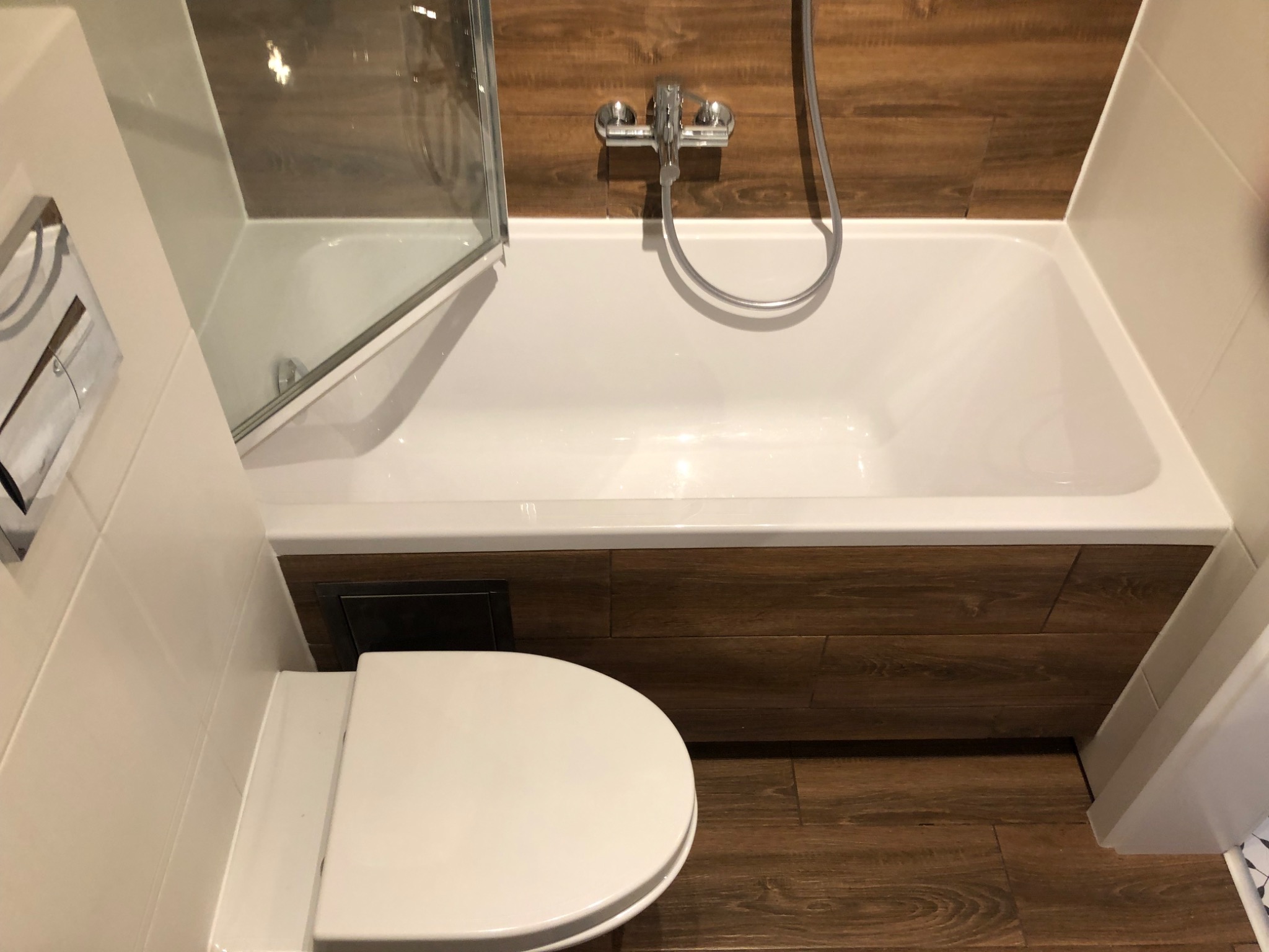 Nowoczesna łazienka z wanną obudowaną drewnopodobnymi płytkami, białą ceramiką i szklanym parawanem nawannowym, widok z góry.