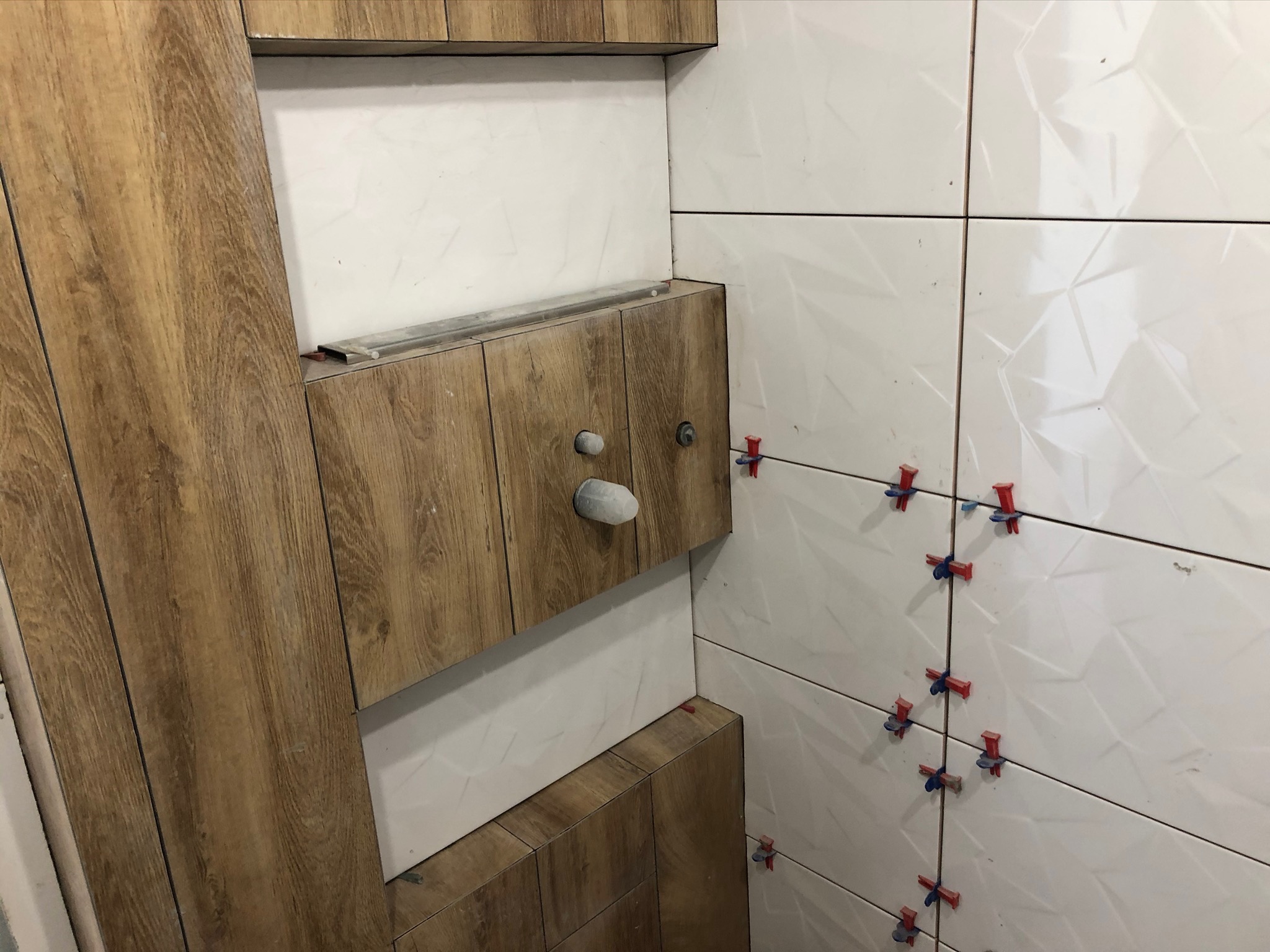 Białe, trójwymiarowe płytki ścienne w trakcie montażu, z użyciem czerwonych krzyżyków dystansowych, sąsiadujące z drewnopodobną zabudową szafki z wystającymi rurami.