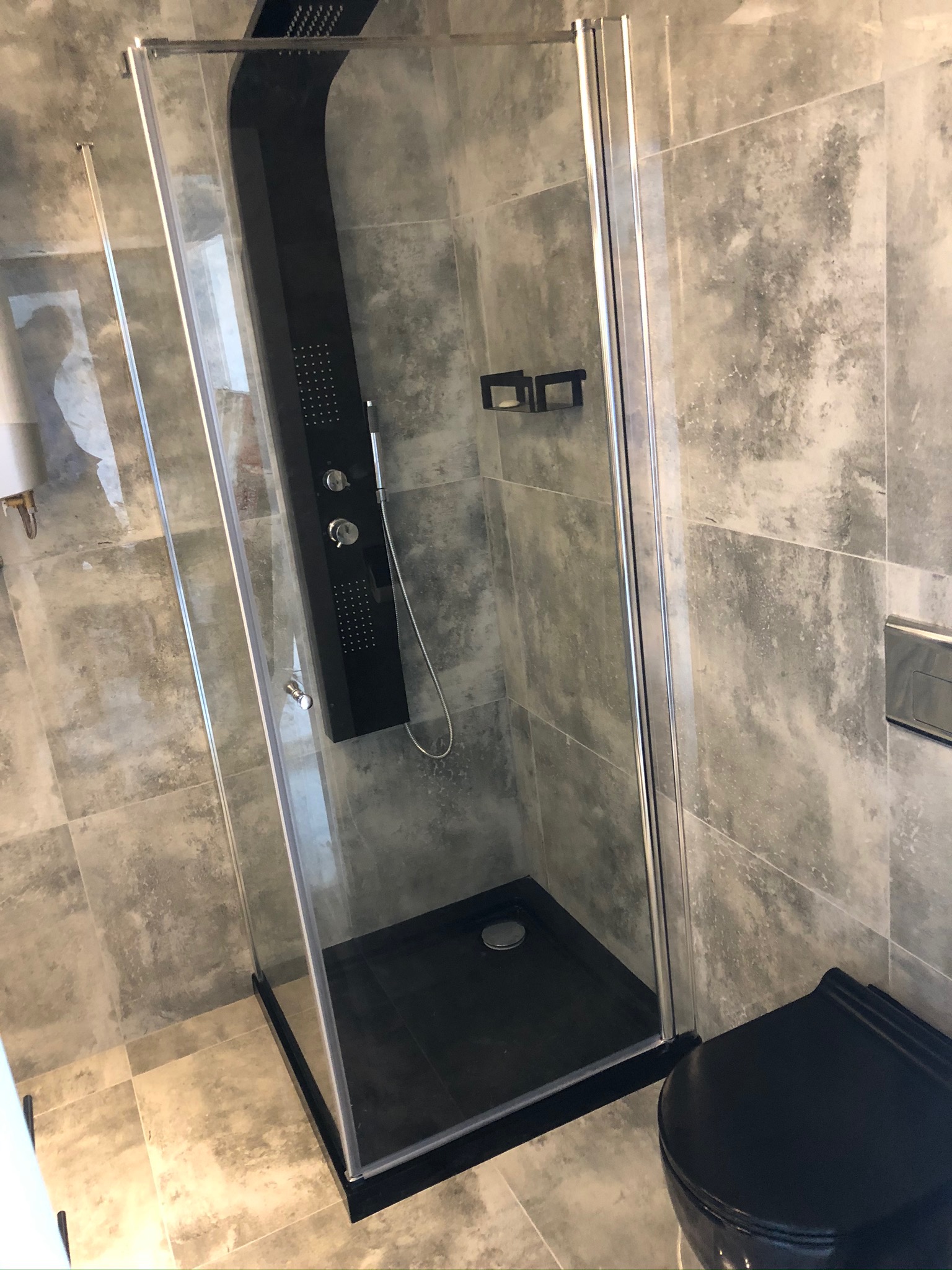 Nowoczesna kabina prysznicowa z czarnym panelem natryskowym i brodzikiem, szare płytki imitujące beton na ścianach i podłodze, widoczny fragment czarnej toalety w rogu.