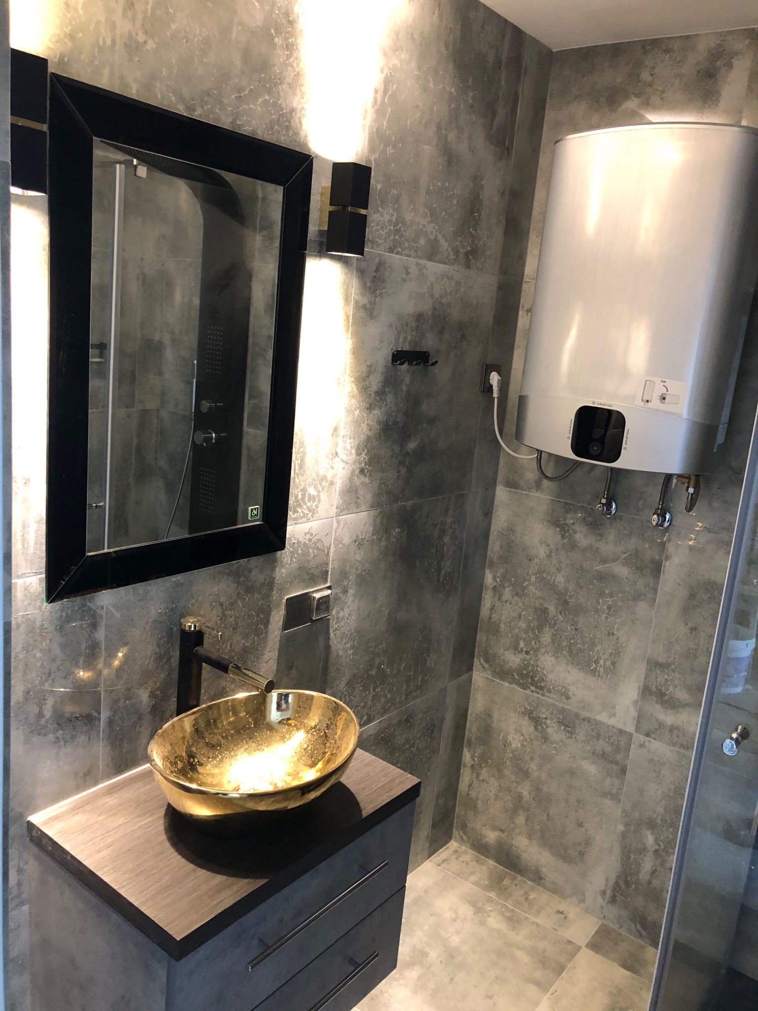 Nowoczesna łazienka z szarymi płytkami imitującymi beton, złotą umywalką nablatową, czarną armaturą, lustrem w czarnej ramie i podgrzewaczem wody.