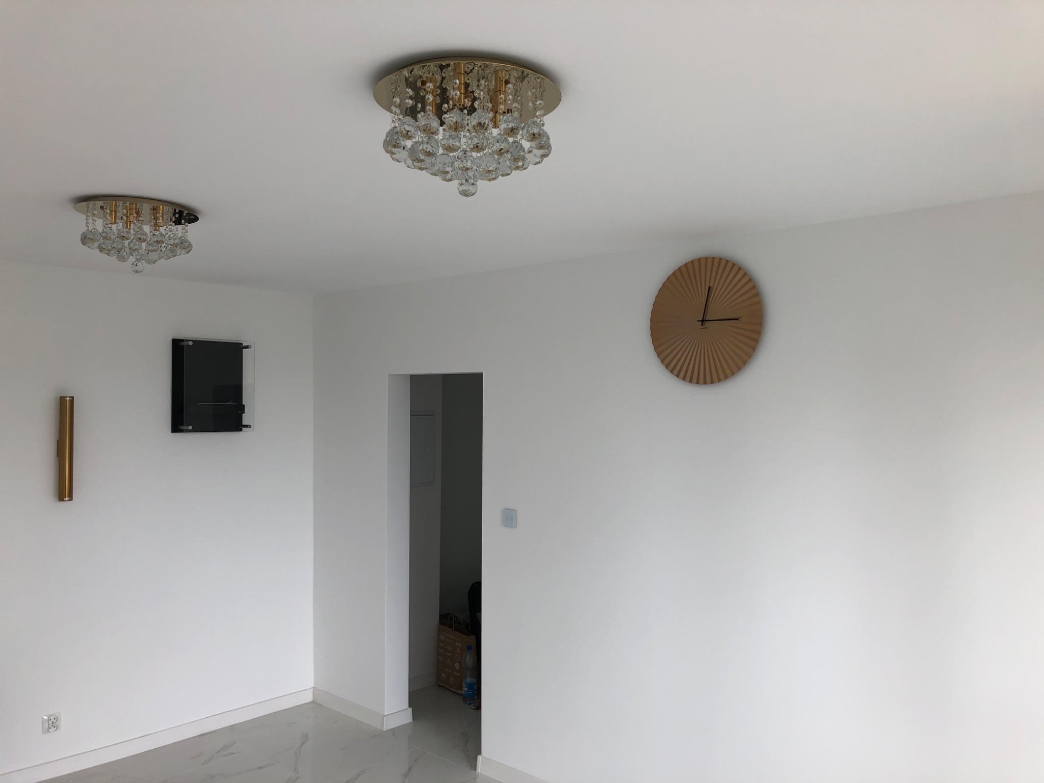 Nowoczesne wnętrze z dwoma kryształowymi lampami sufitowymi, okrągłym drewnianym zegarem na białej ścianie i marmurową podłogą.