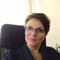 Portret kobiety w okularach i ciemnej marynarce, siedzącej na czarnym krześle biurowym, z widoczną złotą biżuterią na szyi.