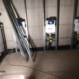 Janusz Dłubak - Montaż stelaży podtynkowych Geberit w łazience w trakcie remontu, widoczne rury kanalizacyjne i profile metalowe.