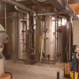 Instalacja przemysłowa z rurami, zbiornikami i zaworami, fotografowana przez instalatora telefonem, obok spawarka i elektryk układający kable.