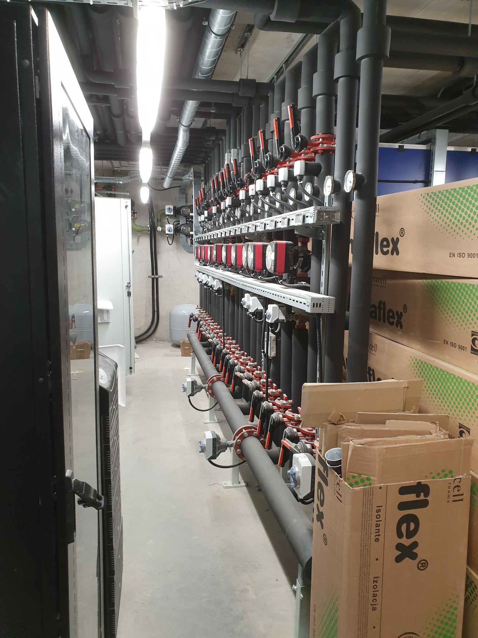 Rozbudowana instalacja grzewcza w kotłowni, widoczne rury z zaworami, manometry, czarne izolacje termiczne oraz kartony z materiałami izolacyjnymi.