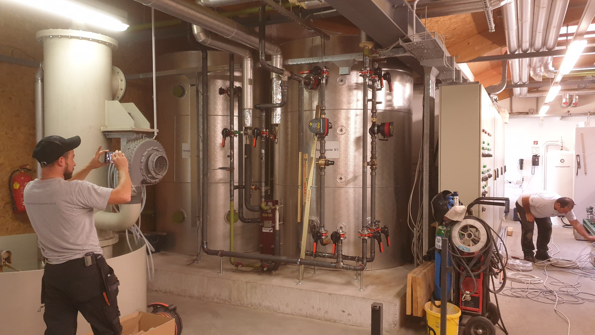 Instalacja przemysłowa z rurami, zbiornikami i zaworami, fotografowana przez instalatora telefonem, obok spawarka i elektryk układający kable.