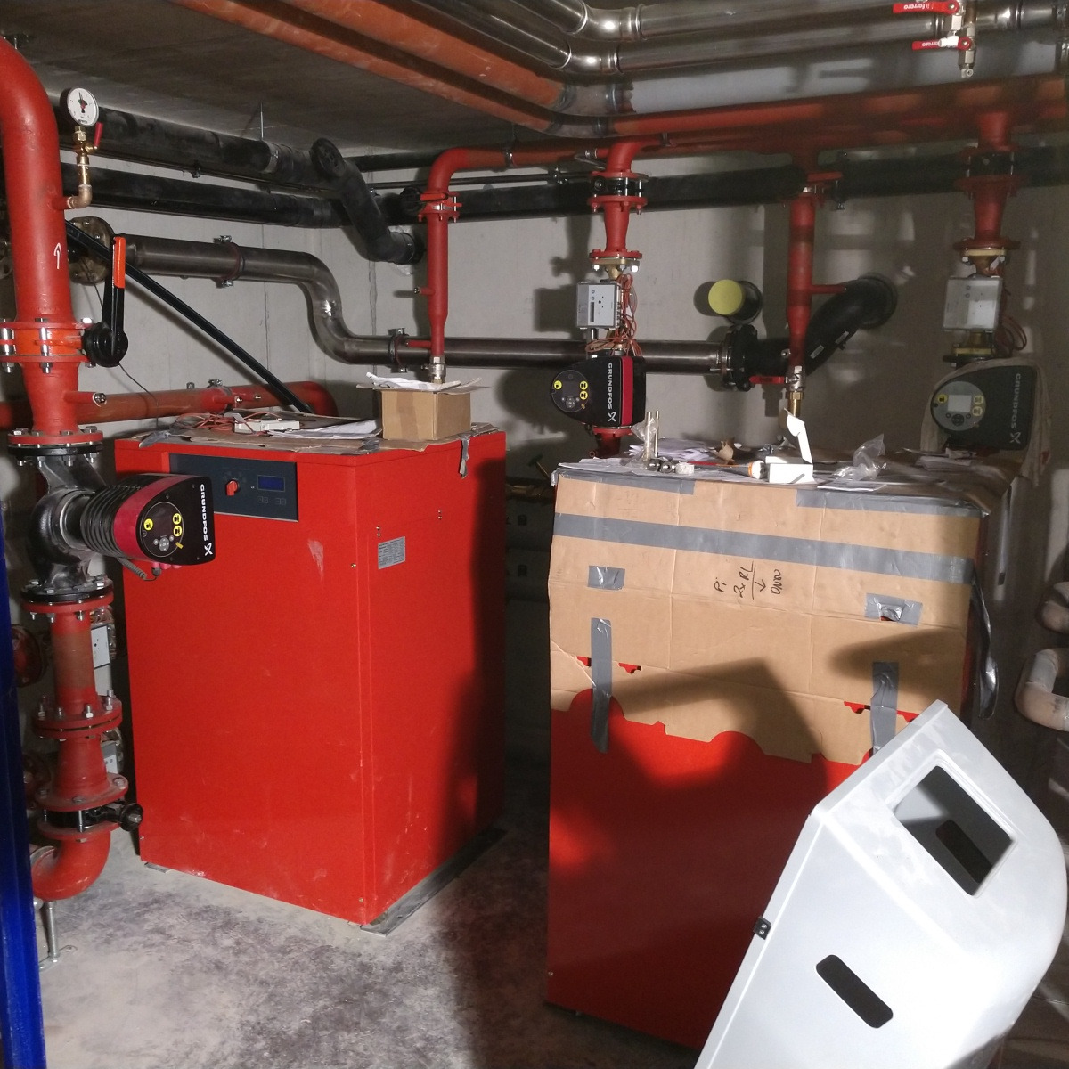 Kotłownia w trakcie montażu: czerwony kocioł grzewczy z podłączonymi rurami i pompami, obok kartonowe opakowanie z taśmą i częściami, betonowa podłoga.