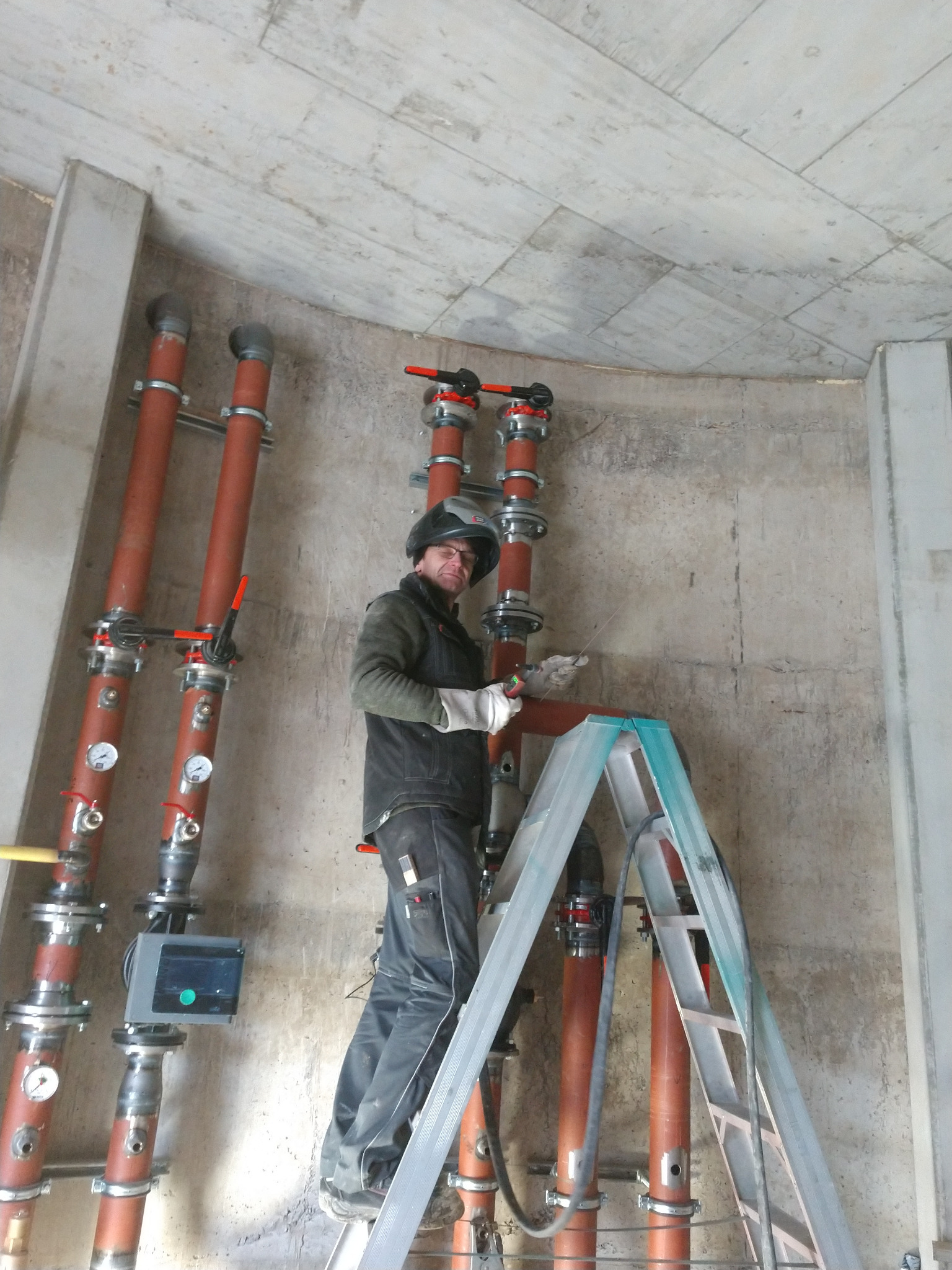 Instalator w hełmie i okularach ochronnych, stojący na aluminiowej drabinie, montuje rury w betonowym pomieszczeniu z widocznymi manometrami i zaworami.