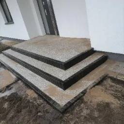 Schody wejściowe do domu wyłożone jasną kostką z ciemnymi obrzeżami, widoczne ślady piasku i ziemi po budowie.