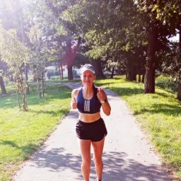 Uśmiechnięta kobieta w sportowym stroju, z daszkiem i zegarkiem na ręce, biegnie ścieżką w parku, pokazując dwa kciuki w górę.