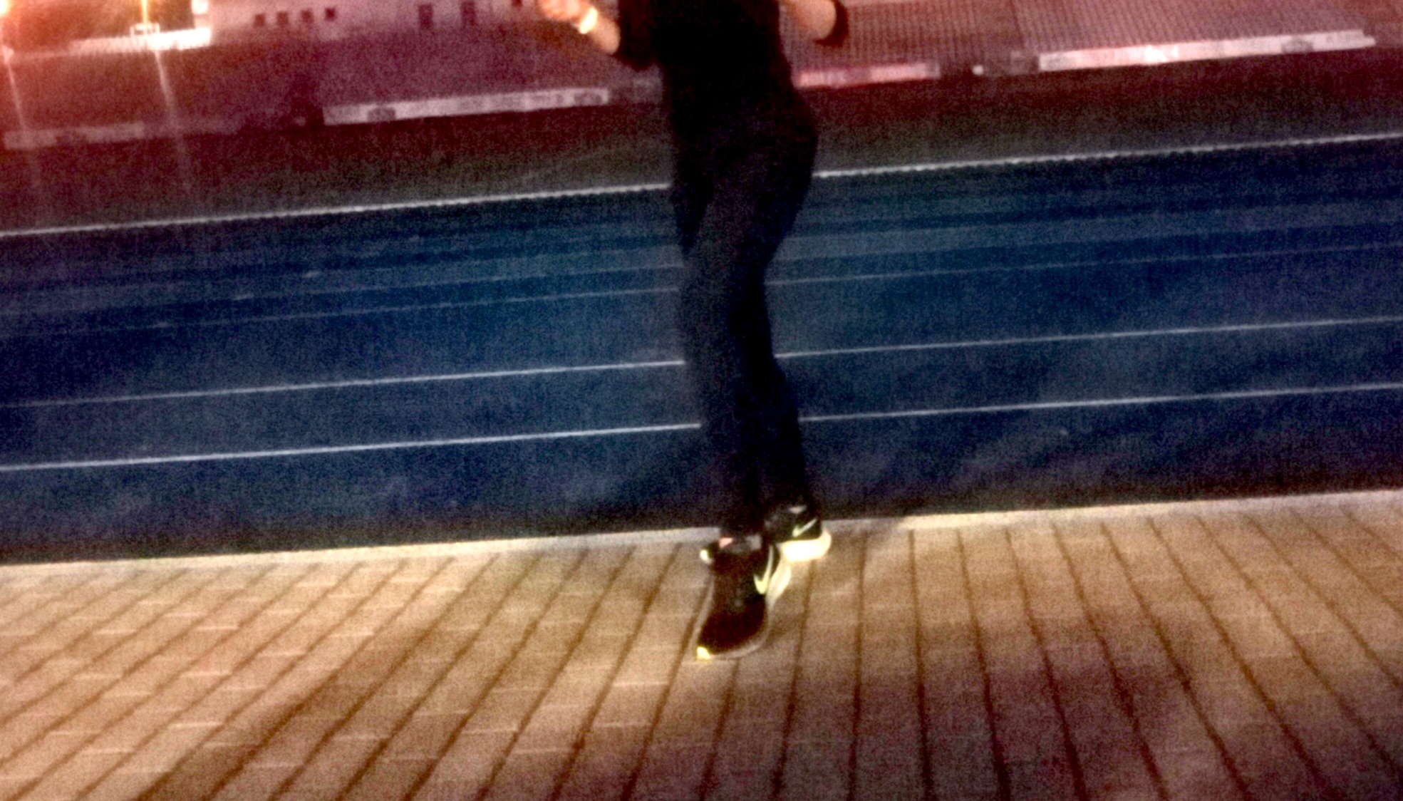 Osoba w sportowym stroju i butach stoi na bieżni tartanowej, widoczne nogi i fragment sylwetki, w tle trybuny.