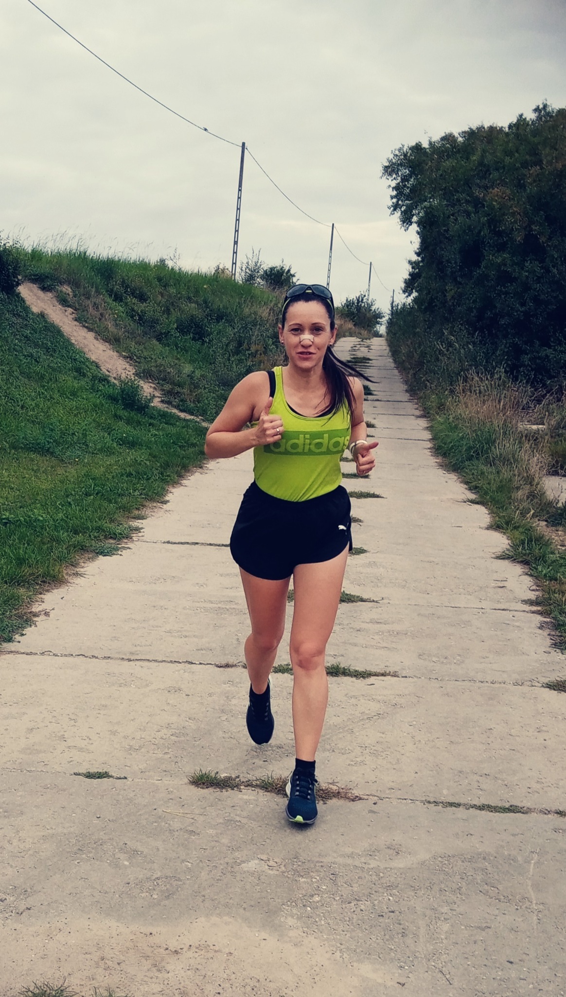 Kobieta w sportowym stroju, z plastrem na nosie, biegnie pod górę betonową ścieżką, kciuki uniesione w górę.