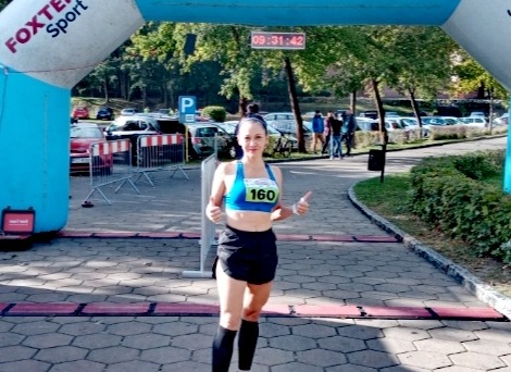 Uśmiechnięta kobieta z numerem startowym 160, w sportowym stroju, pokazuje kciuki w górę na mecie biegu; w tle brama startowa Foxtel Sport i zaparkowane samochody.