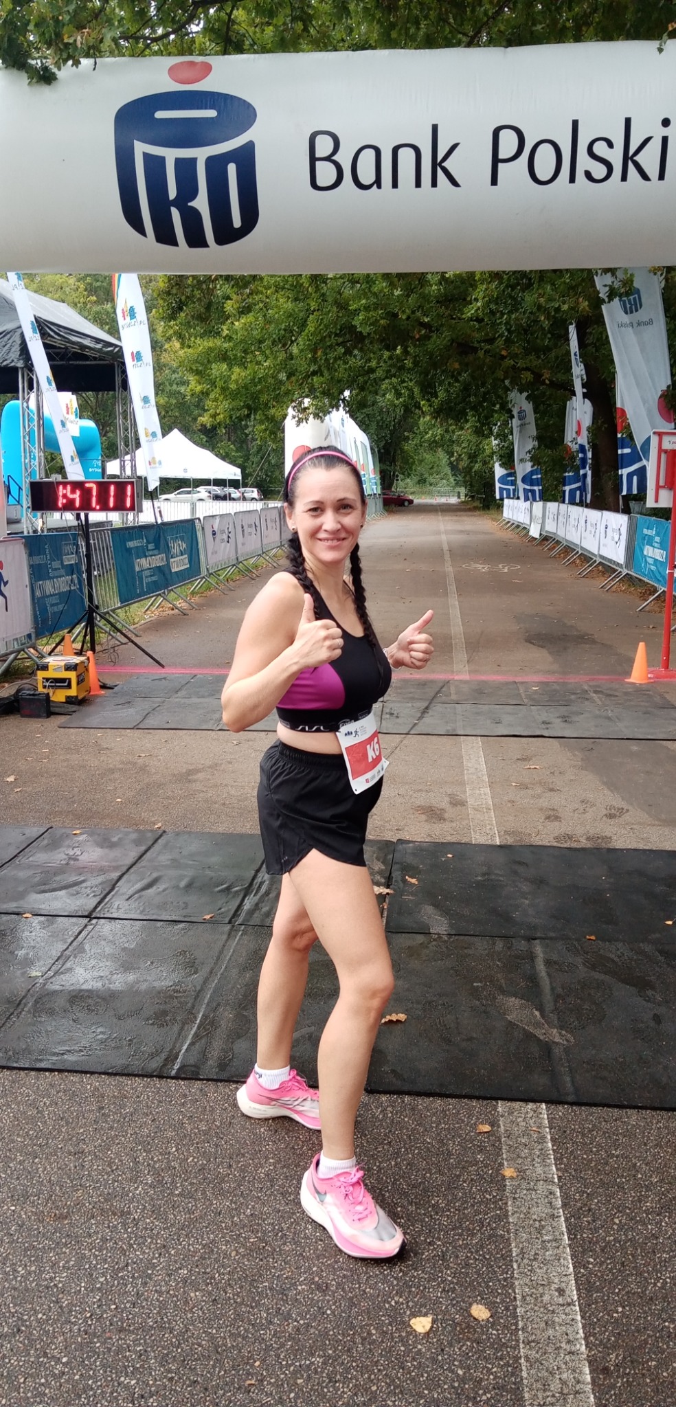 Uśmiechnięta kobieta z numerem startowym K6 na mecie biegu, podnosi kciuki w górę, za nią baner Banku Polskiego i tablica z czasem 1:47:11.