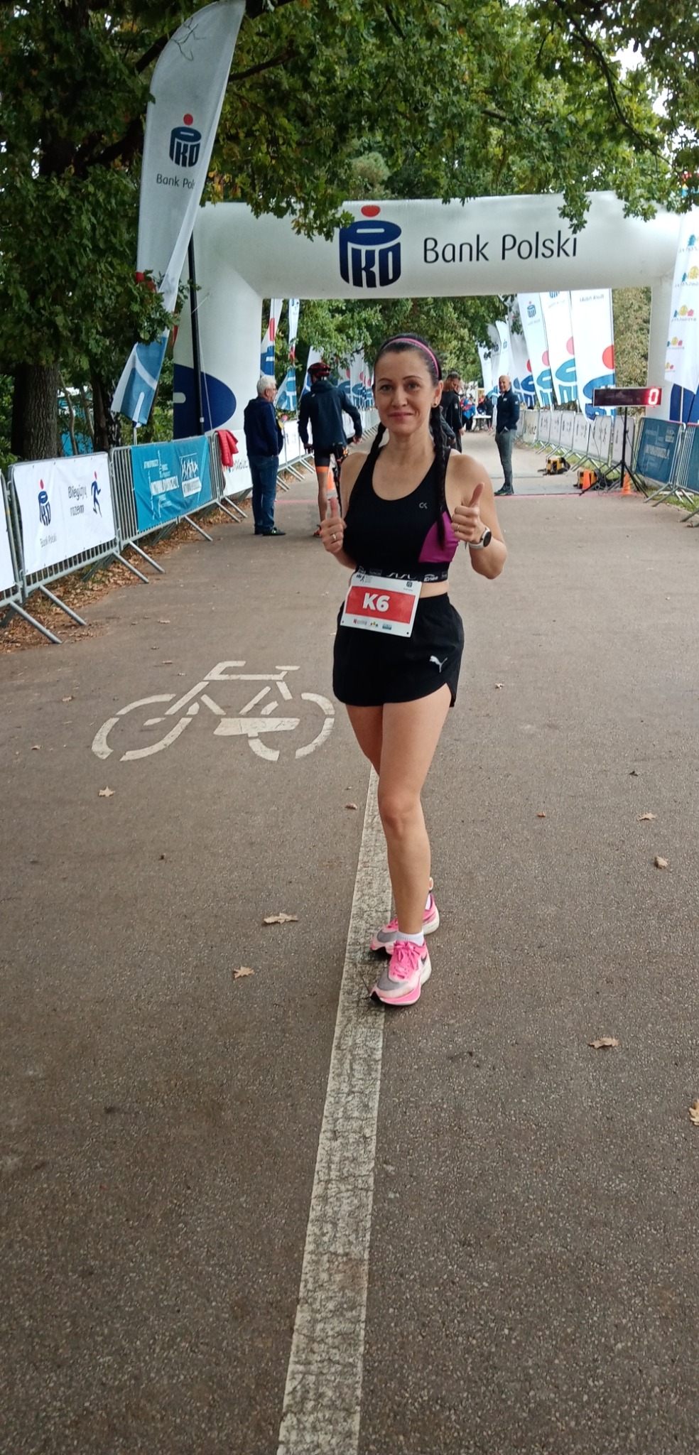 Uśmiechnięta kobieta w sportowym stroju, z numerem startowym K6, pokazuje kciuki w górę na tle mety biegu ulicznego z logo sponsora Bank Polski.