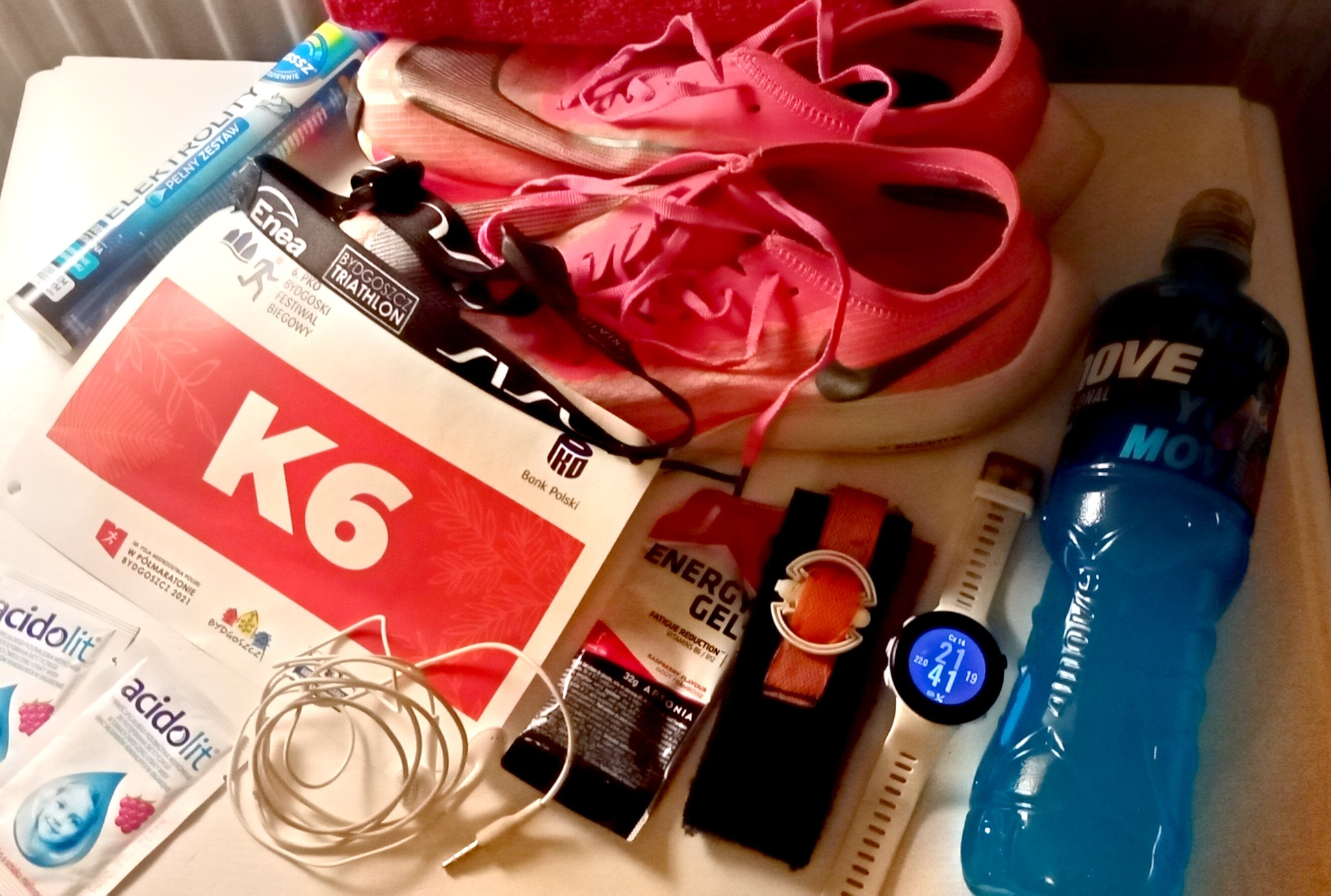 Różowe buty do biegania, bidon z niebieskim napojem izotonicznym, żel energetyczny, zegarek sportowy, elektrolity i pakiet startowy na Bydgoszcz Triathlon K6.