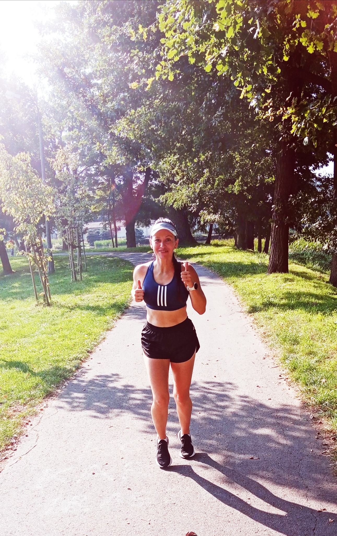 Uśmiechnięta kobieta w sportowym stroju, z daszkiem i zegarkiem na ręce, biegnie ścieżką w parku, pokazując dwa kciuki w górę.