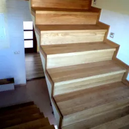 Jasne, drewniane schody z widocznym usłojeniem, prowadzące w górę, z widoczną dalszą częścią schodów w tle oraz fragmentem ściany z włącznikiem.