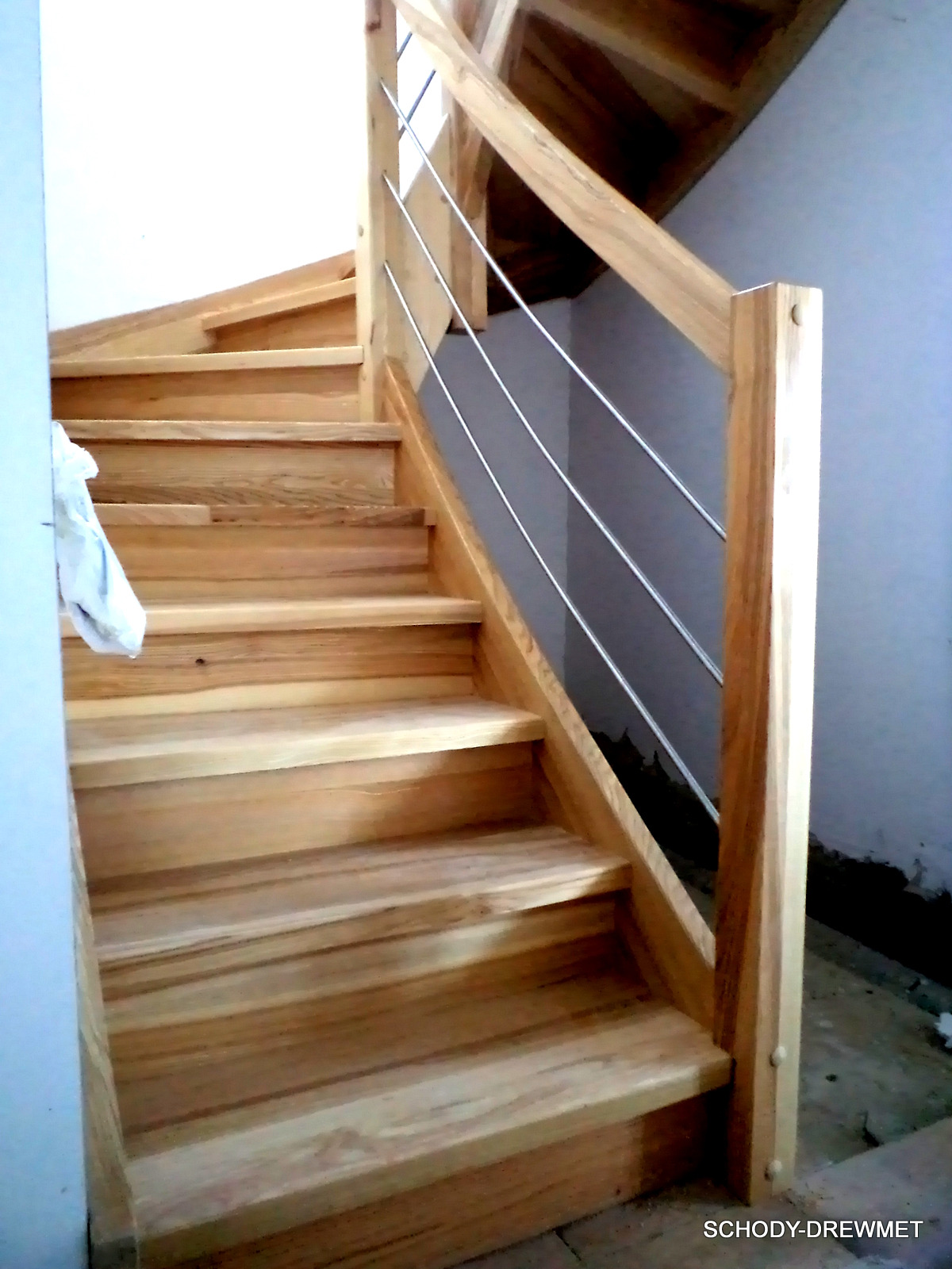 Jasne, drewniane schody z metalową balustradą w Regulicach, widziane z dołu, z fragmentem białej ściany po lewej stronie i surowym wykończeniem podłogi po prawej.