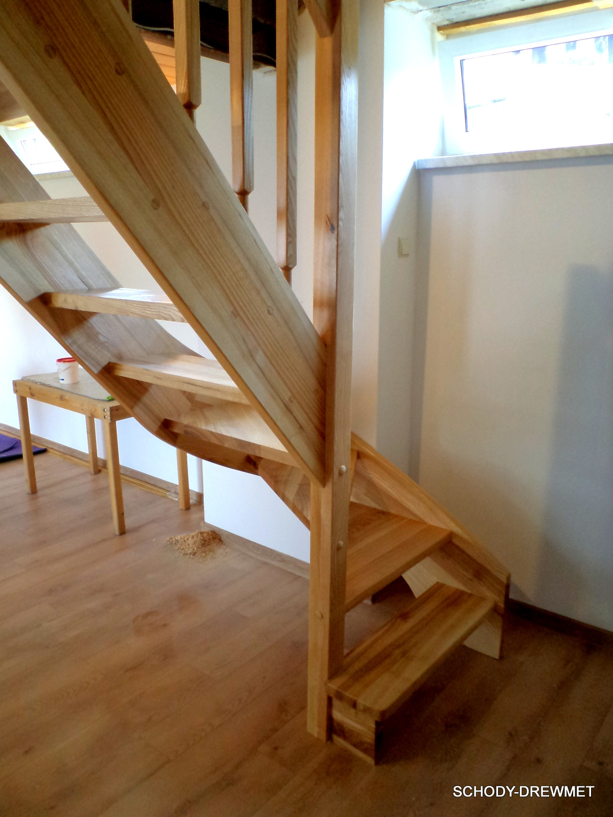 Jasne, sosnowe stopnie schodów z widocznym usłojeniem, na podłodze leżą wióry, w tle biała ściana i okno.