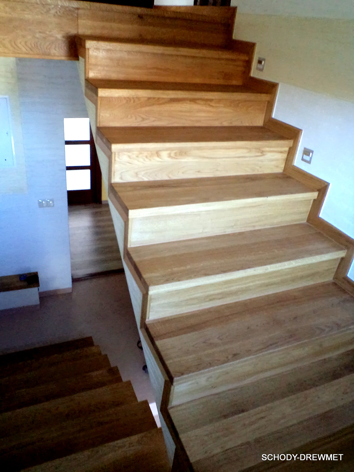 Jasne, drewniane schody z widocznym usłojeniem, prowadzące w górę, z widoczną dalszą częścią schodów w tle oraz fragmentem ściany z włącznikiem.