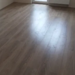 Ułożone panele podłogowe w jasnym odcieniu dębu z widocznymi śladami stóp, świeżo po montażu, w nowym pokoju z białymi ścianami i grzejnikiem.