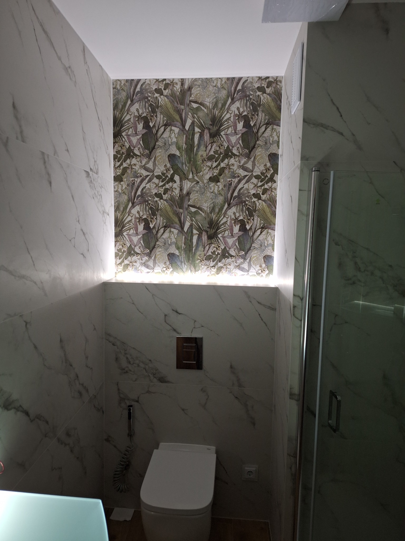 Nowoczesna łazienka z marmurowymi płytkami, toaleta podwieszana z chromowanym przyciskiem, tropikalna tapeta podświetlona ledami i kabina prysznicowa.