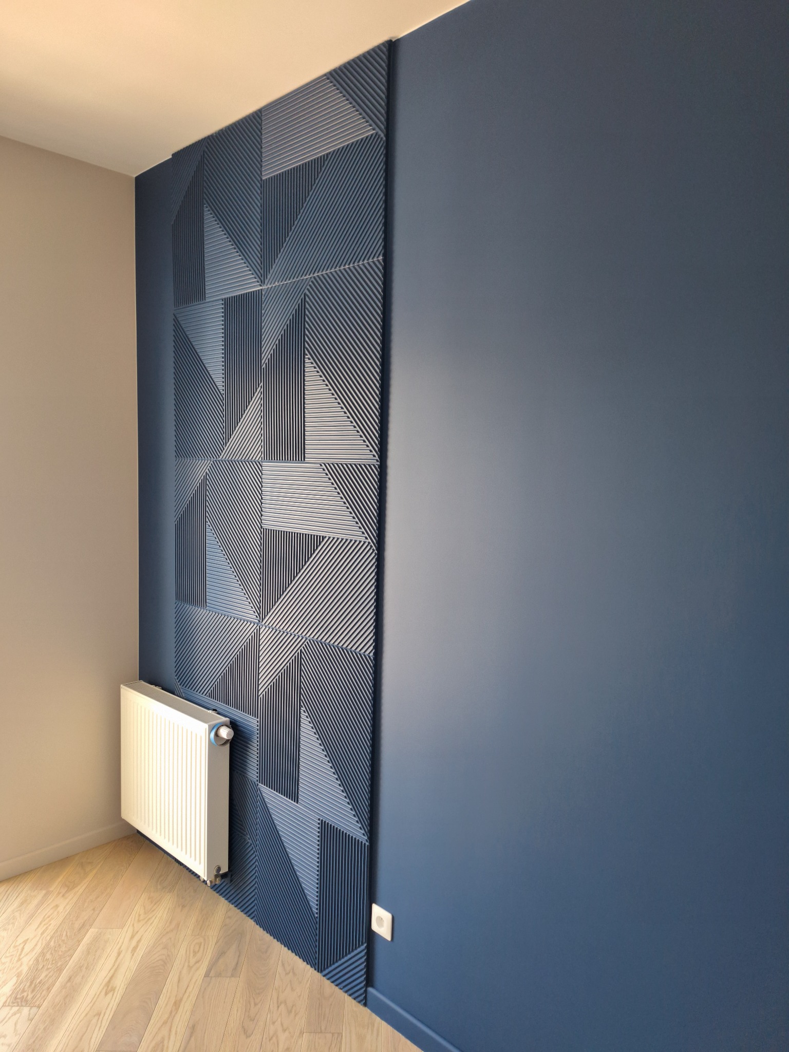 Granatowa ściana z geometrycznym panelem dekoracyjnym, biały grzejnik na tle drewnianej podłogi.