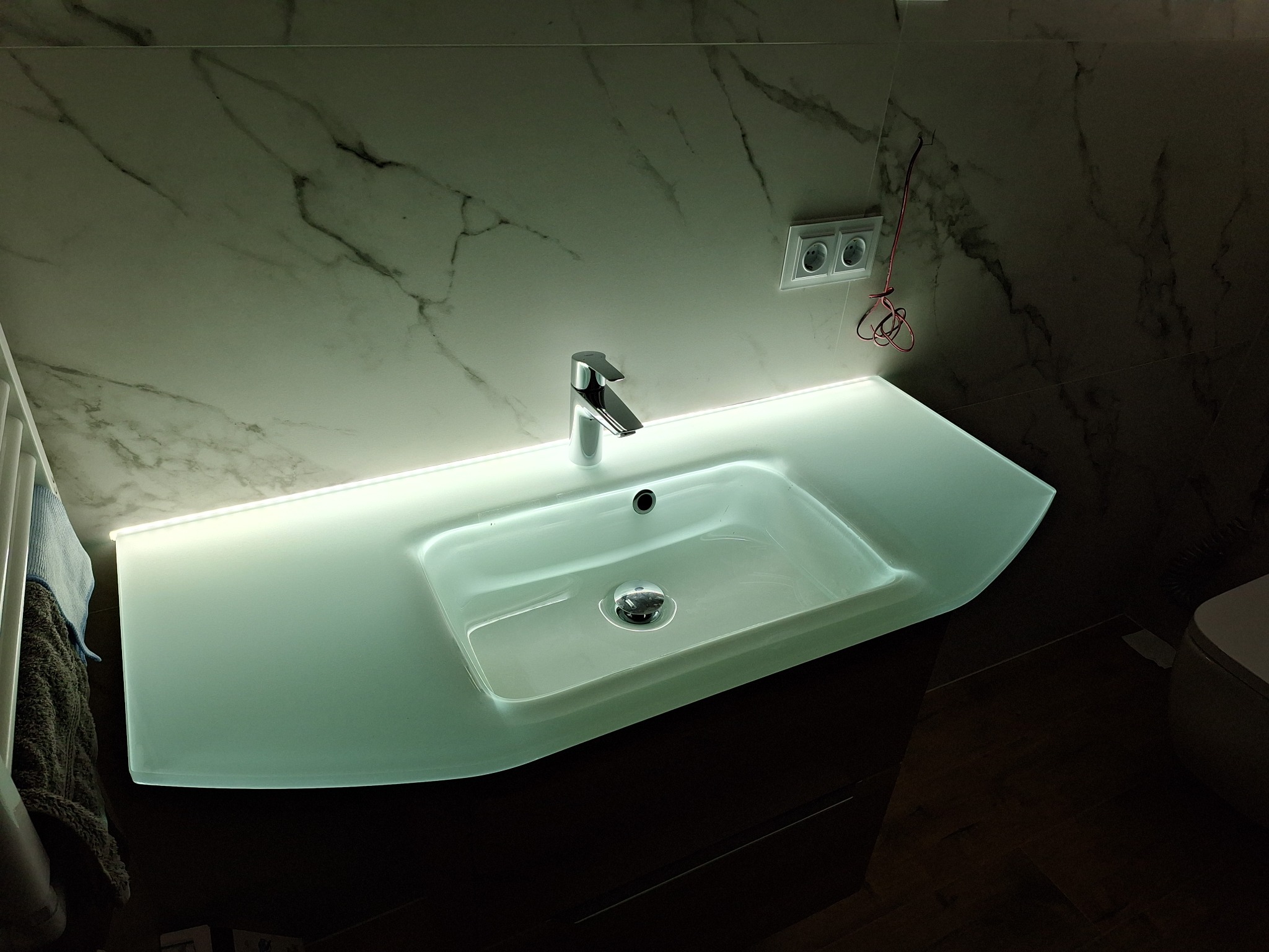 Nowoczesna umywalka z podświetleniem LED, bateria chromowana, marmurowe płytki na ścianie, widoczne gniazdka elektryczne i fragment toalety.