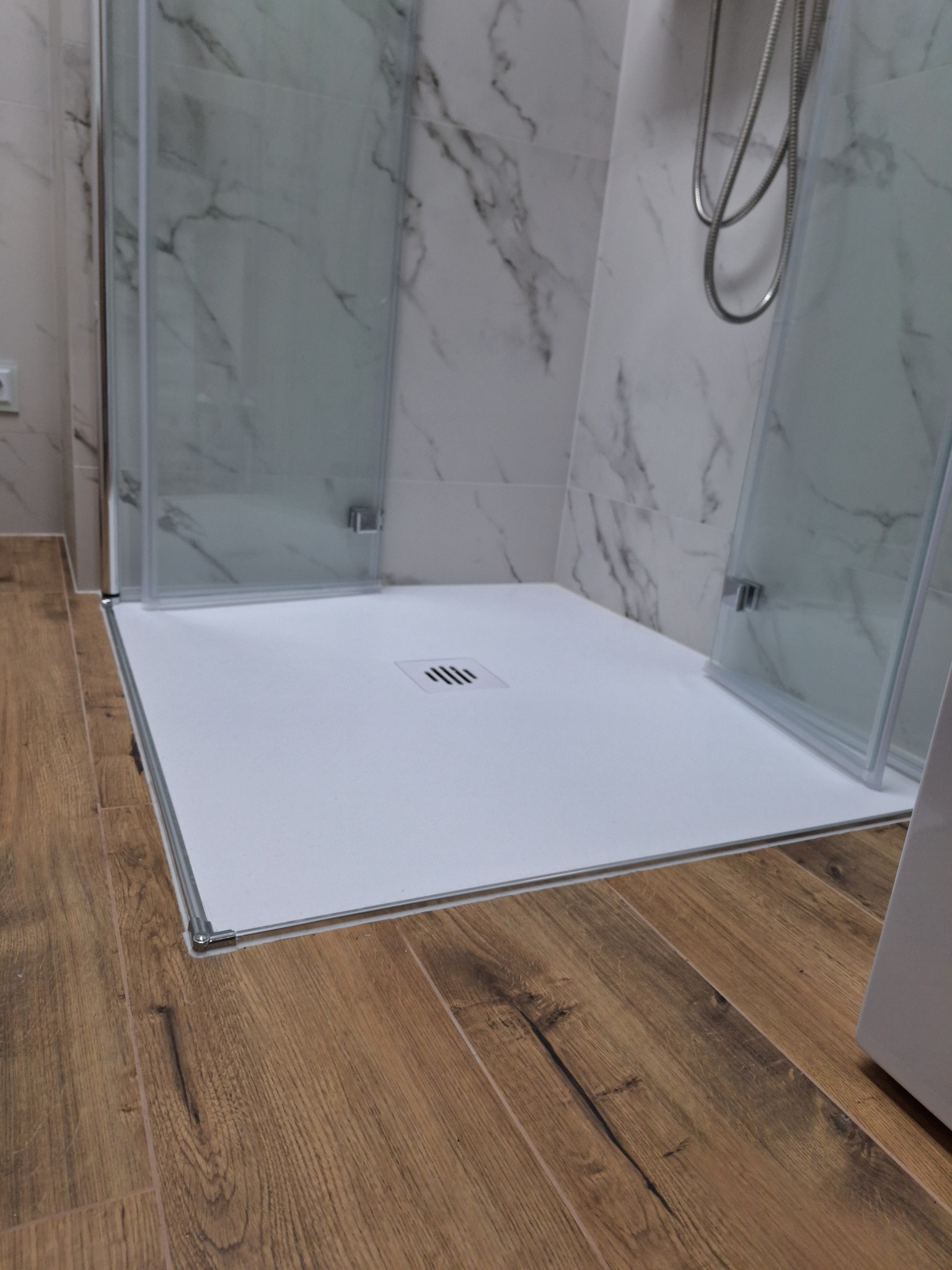 Nowoczesny brodzik z odpływem liniowym, kabina prysznicowa z przezroczystego szkła, marmurowe płytki na ścianie i drewnopodobna podłoga.