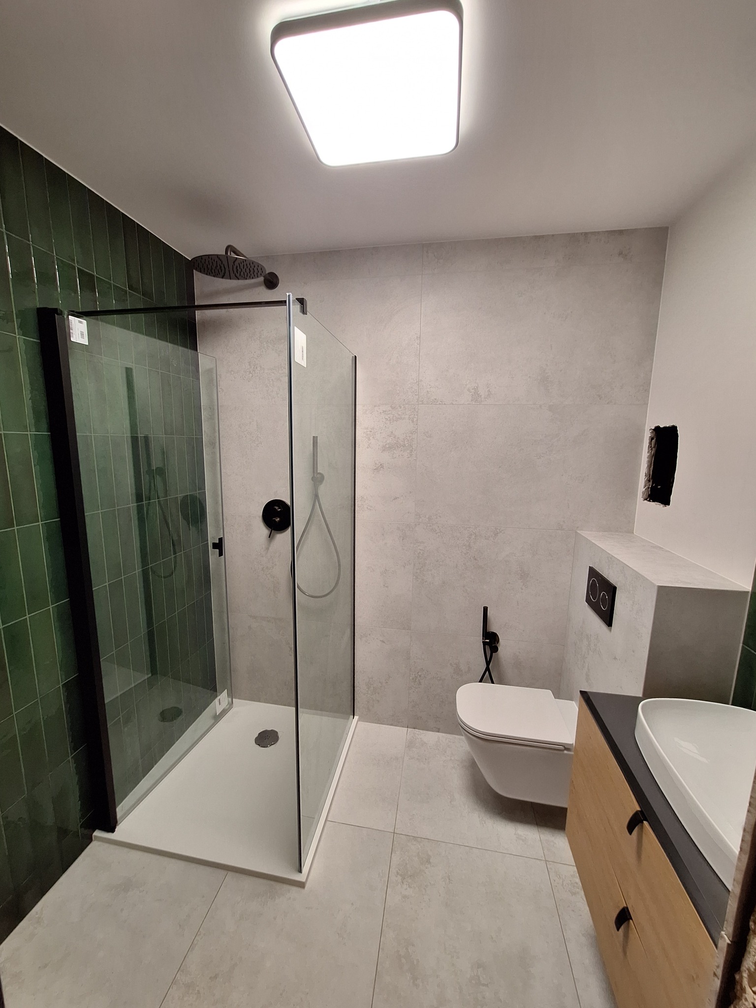 Nowoczesna łazienka z kabiną typu walk-in, zielonymi płytkami i minimalistyczną armaturą w czarnym kolorze. Umywalka z szafką z jasnego drewna.