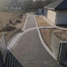 Ukończony podjazd z szarej kostki betonowej, obrzeża z jasnej kostki, widoczne domy w tle i brama wjazdowa rzucająca cień na nawierzchnię.