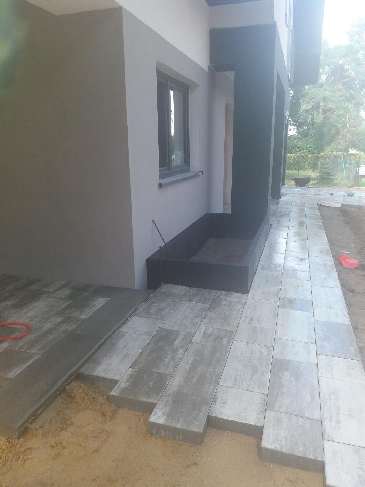 Fragment układanej nawierzchni z prostokątnych, szarych elementów na podsypce piaskowej, widoczne krawężniki i budynek w tle.