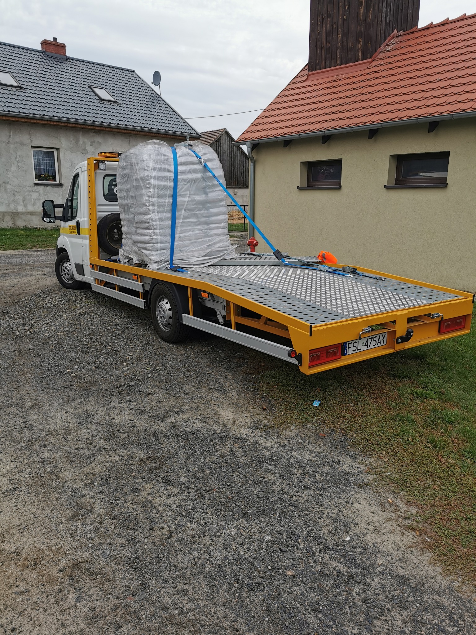 Żółta platforma samochodu pomocy drogowej przewozi zabezpieczony ładunek owinięty folią, w tle budynki mieszkalne.