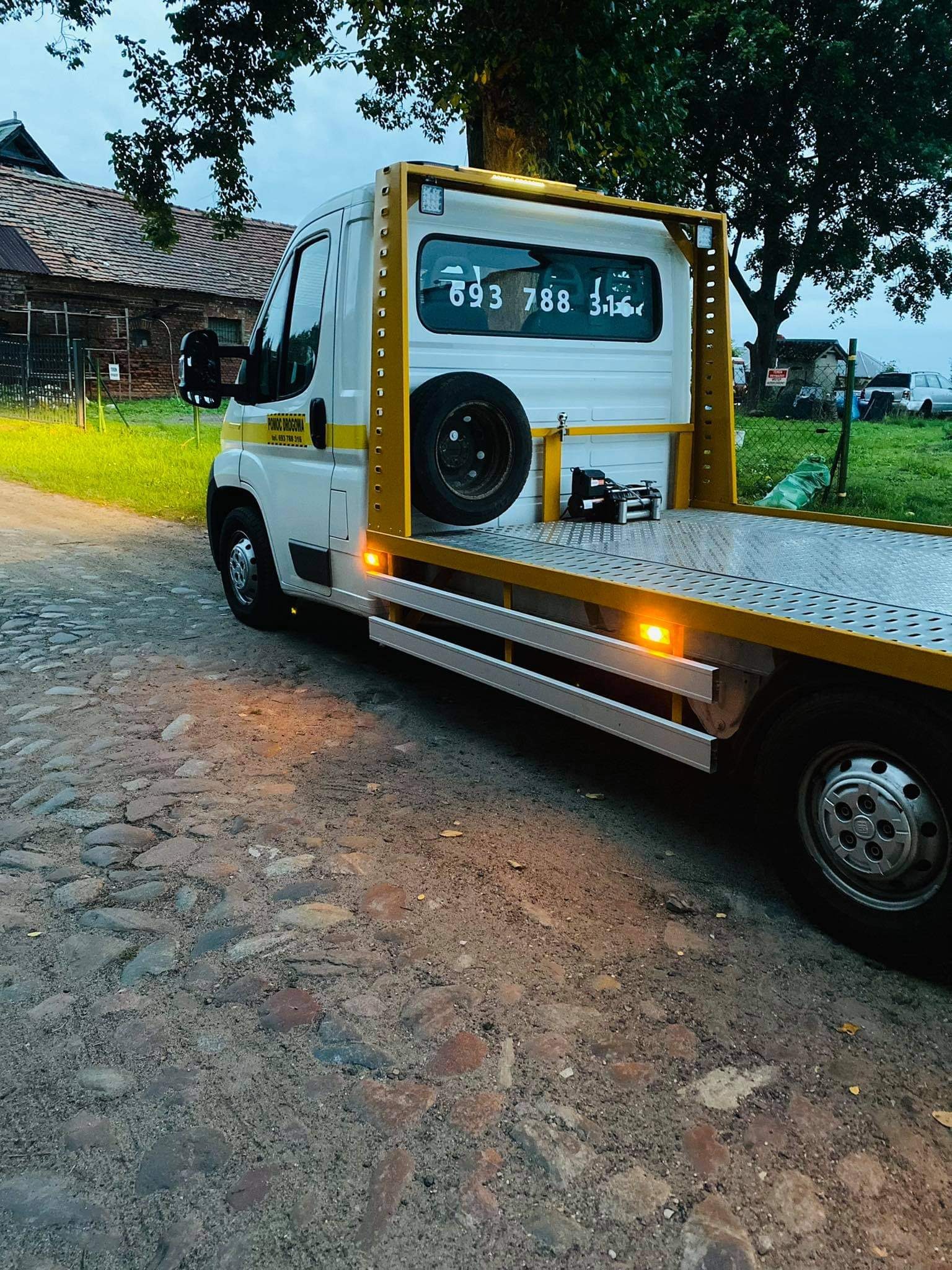 Biała laweta z żółtą ramą na brukowanej drodze, widoczny numer telefonu na szybie i koło zapasowe.