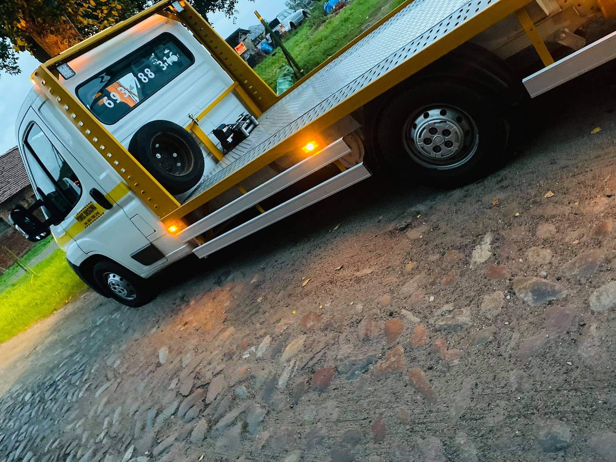 Biała laweta z żółtymi elementami konstrukcyjnymi, zaparkowana na brukowanej drodze, widoczny numer telefonu na szybie.