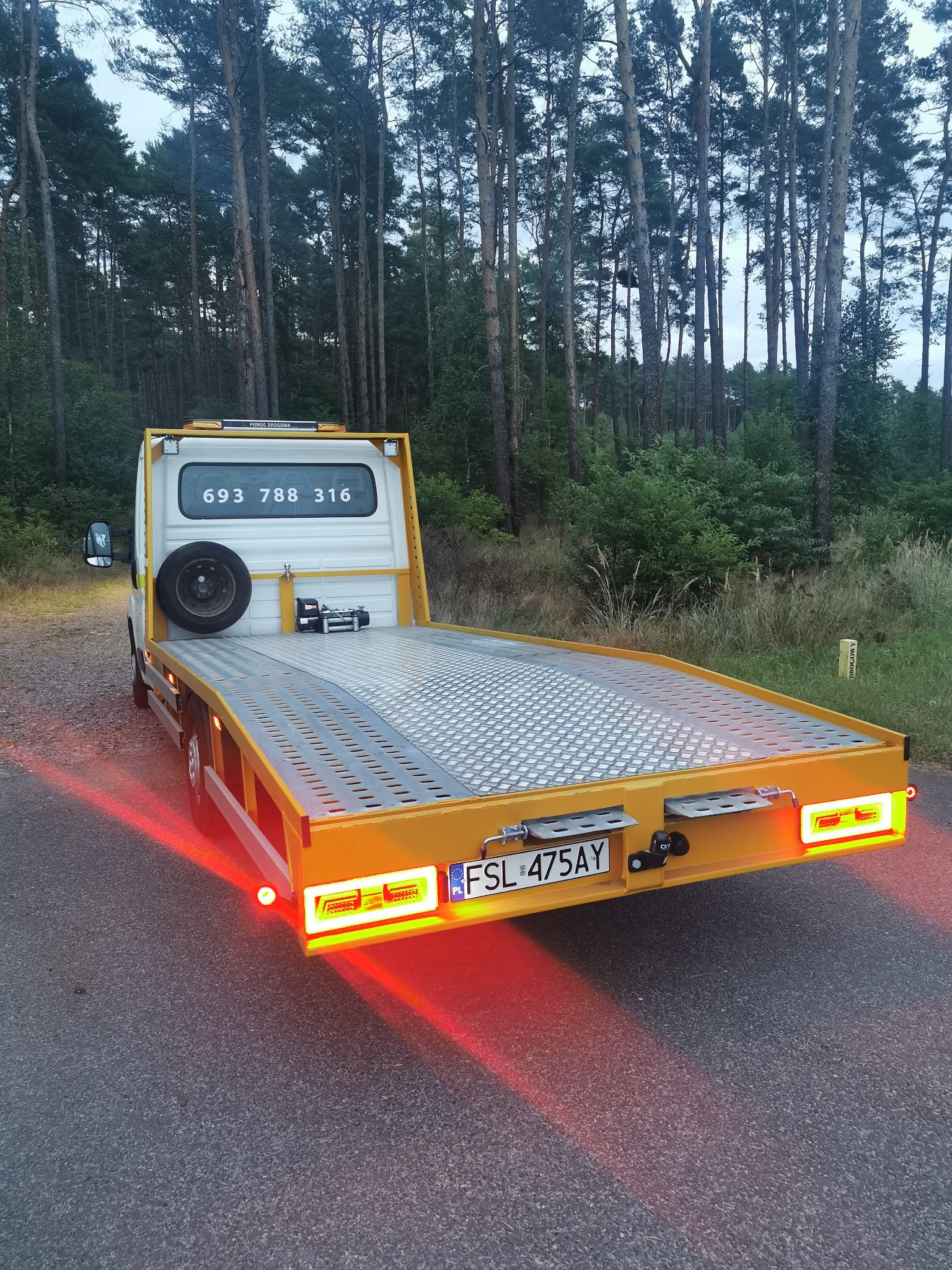 Żółta laweta z numerem telefonu na tle leśnym, widziana z tyłu, światła stopu włączone, pusta platforma załadunkowa