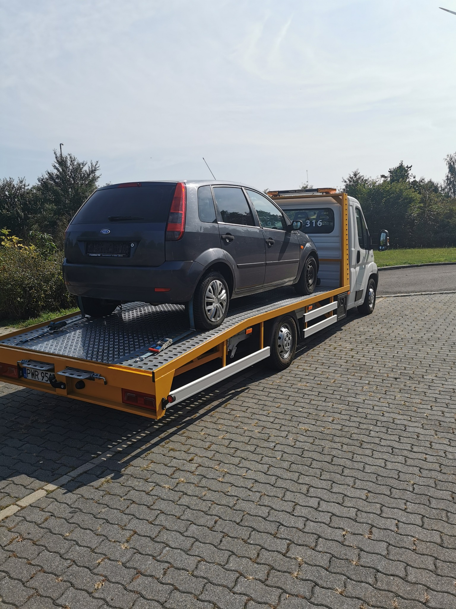 Szary Ford Fiesta przewożony na żółtej lawecie z numerem 316 na tle brukowanej powierzchni i zieleni.