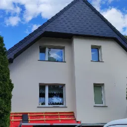 Dwupiętrowy dom z nowym, ciemnym dachem krytym dachówką. Widoczne rusztowanie i przygotowania do prac elewacyjnych. Bujna zieleń drzew w tle.