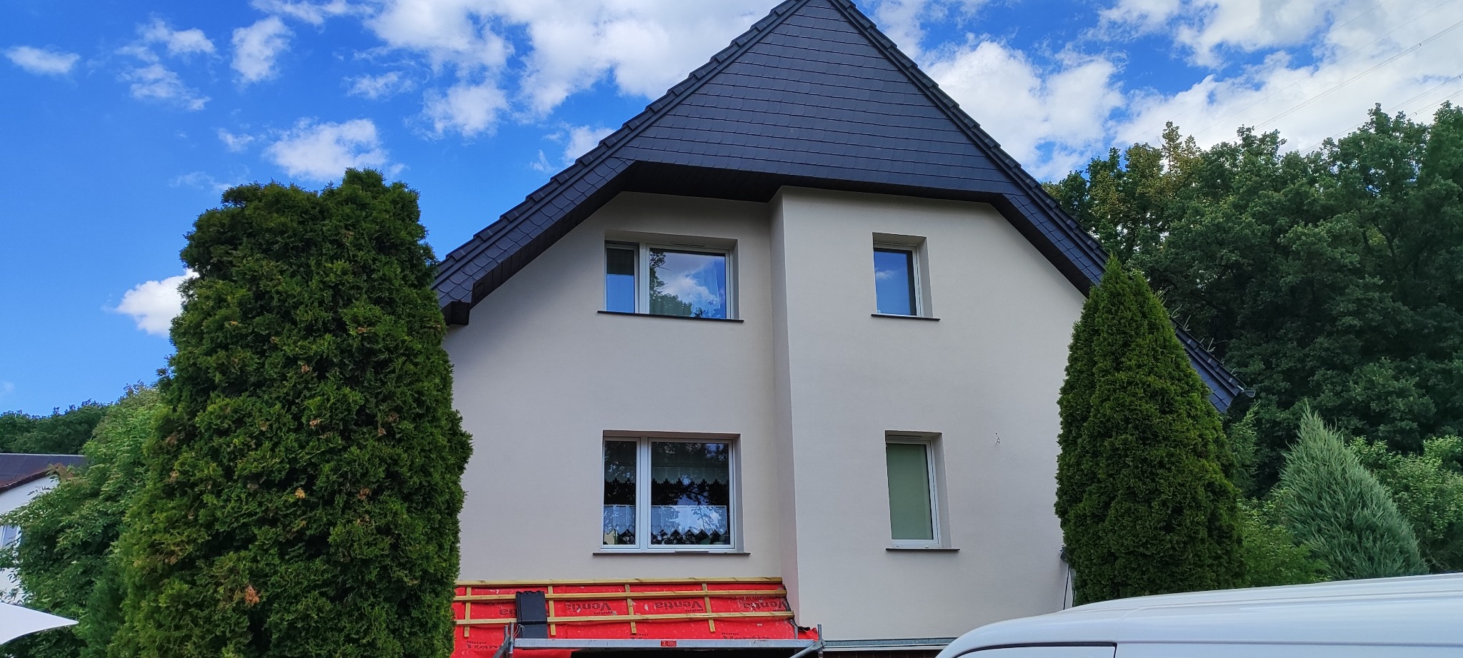 Dwupiętrowy dom z nowym, ciemnym dachem krytym dachówką. Widoczne rusztowanie i przygotowania do prac elewacyjnych. Bujna zieleń drzew w tle.