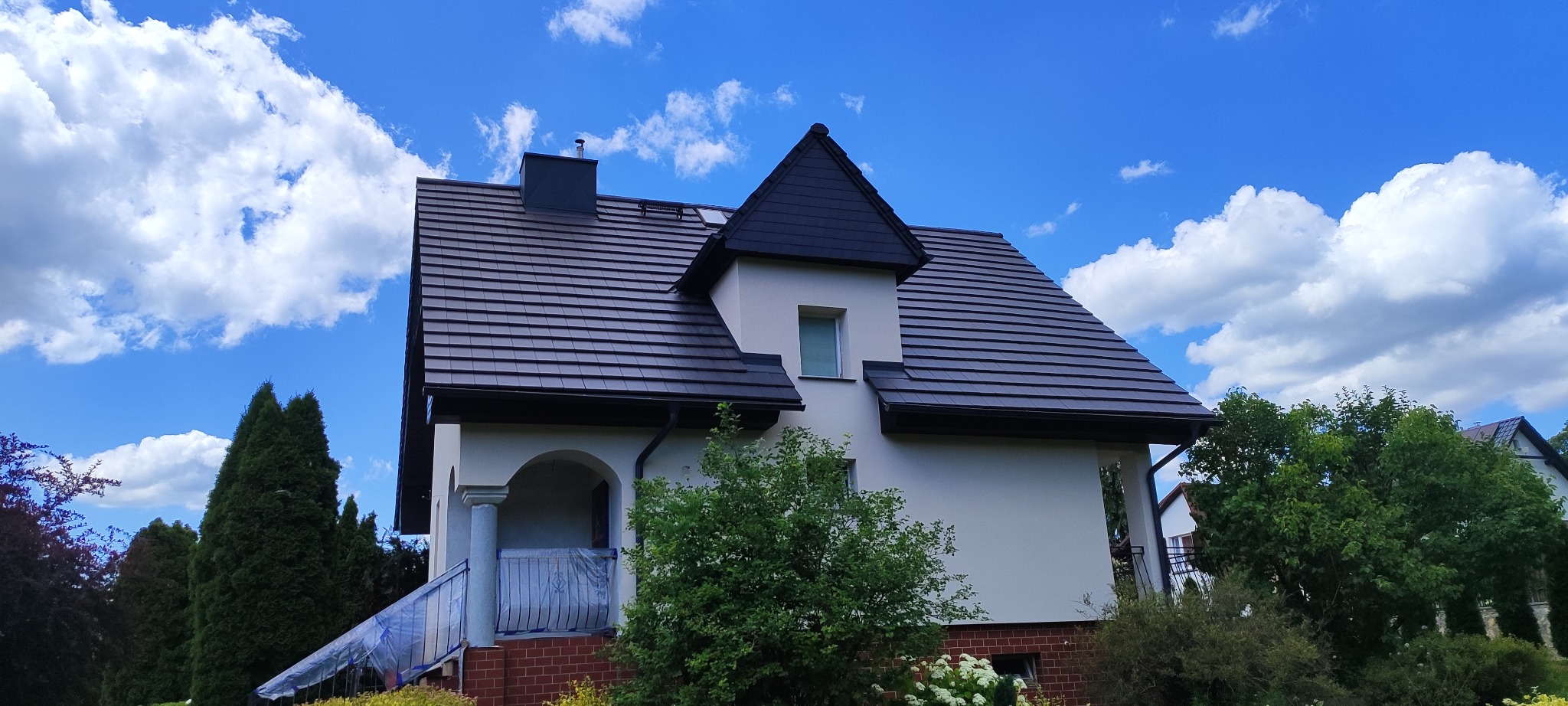 Dom jednorodzinny z ciemną dachówką i jasną elewacją w słoneczny dzień. Widoczny fragment balkonu zabezpieczony folią. Ujęcie z dołu, podkreślające bryłę budynku.