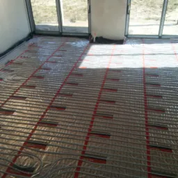 Rozłożone rury ogrzewania podłogowego na folii z siatką w Olsztynie, przygotowane do zalania wylewką, widoczne okna i drzwi balkonowe.